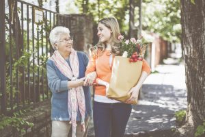 Elderley lady arm in arm with careworker carrying a bunch of flowers on a sunny tree lined urban sidewalk